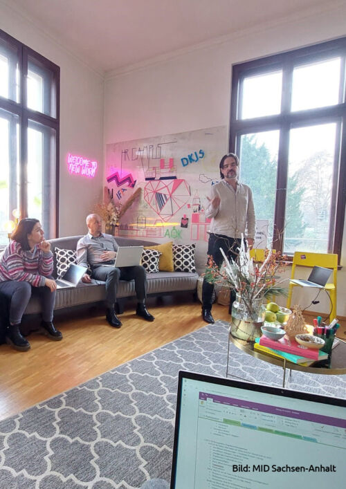Projekttreffen im Zuge der ebenenübergreifenden Projektarbeit. Zu sehen sind drei Personen in einer kreativen Arbeitsumgebung. Bunte Möbel, buntes Arbeitsmaterial. Im Vordergrund ist ein Laptopbildschirm zu sehen, im Hintergrund ein großer Bildschirm, auf dem Inhalte präsentiert werden.