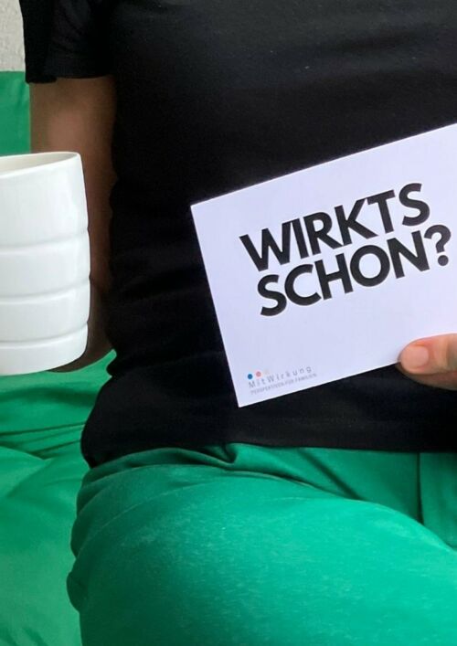 Bild einer sitzender Frau, in der einen Hand eine Kaffeetasse, in der anderen Hand eine Karte mit dem Text: Wirkt's schon?